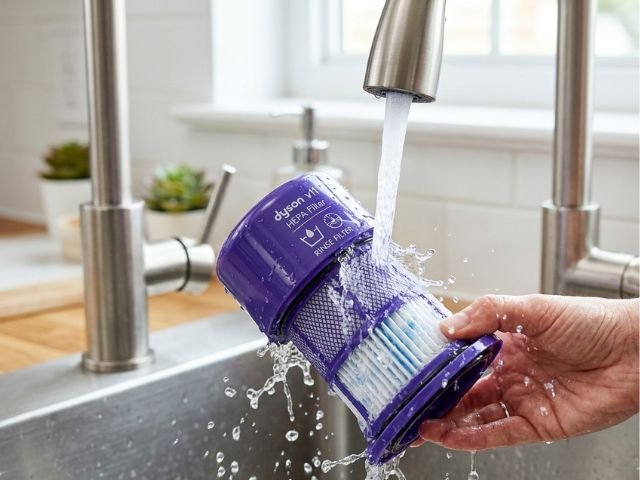 Washing a purple cylindrical vacuum filter under running kitchen tap water to prevent getting a dyson v11 filter message after washing